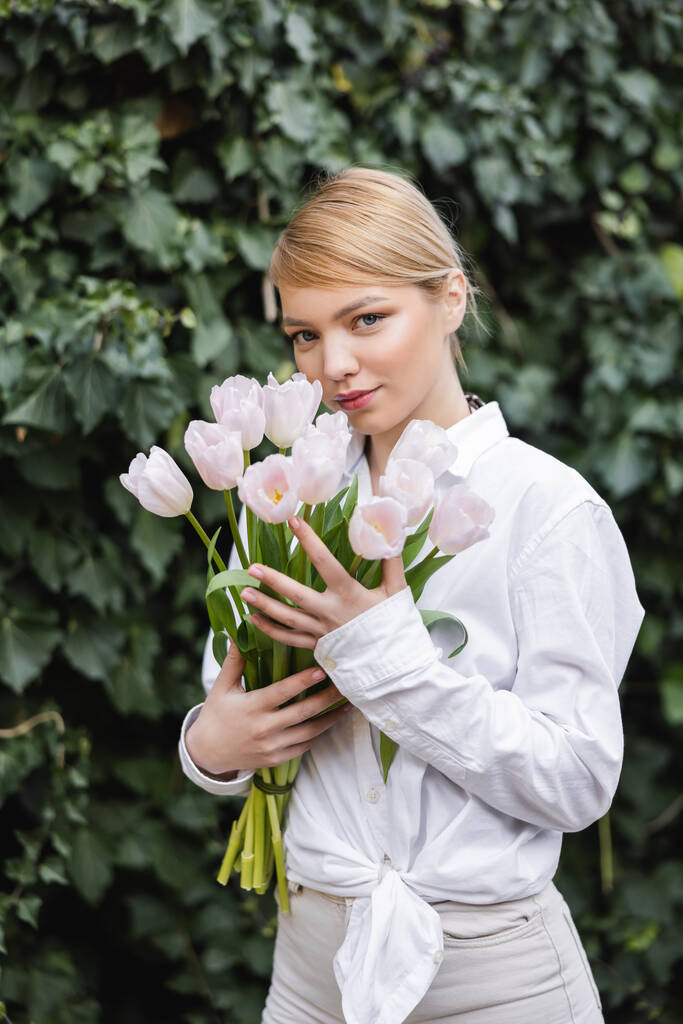 blonde woman in white shirt holding white tulips and looking at camera near green ivy - Photo, Image