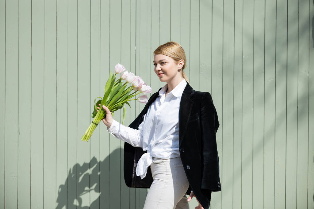 молодая женщина в белой рубашке и черном блейзере идет с тюльпанами возле серой стены - Фото, изображение