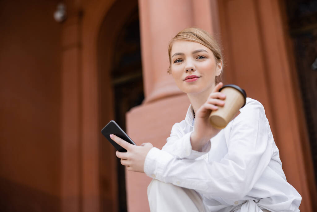 блондинка з паперовою чашкою і мобільним телефоном дивиться на камеру на відкритому повітрі
 - Фото, зображення