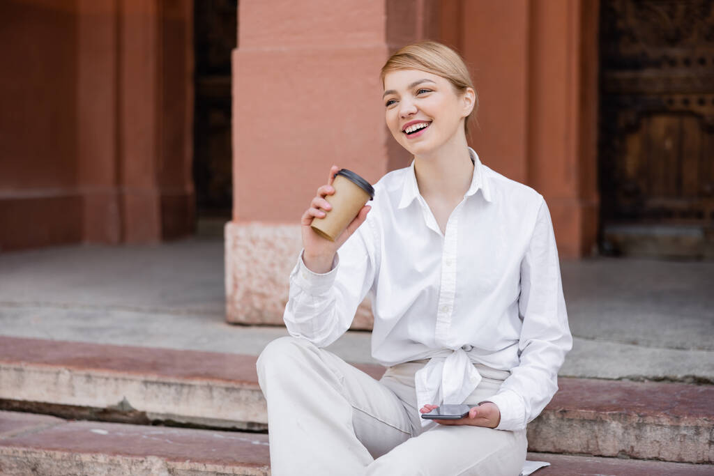 iloinen nuori nainen istuu portaissa matkapuhelimen ja kahvin kanssa mennä - Valokuva, kuva