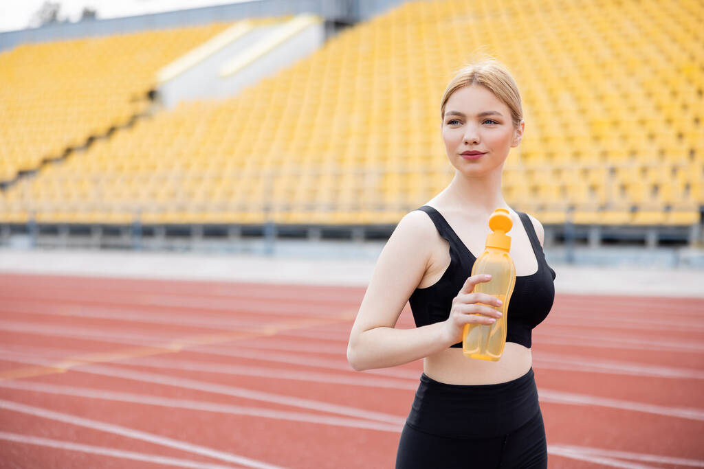 sportovní žena dívá pryč, zatímco drží sportovní láhev na stadionu - Fotografie, Obrázek