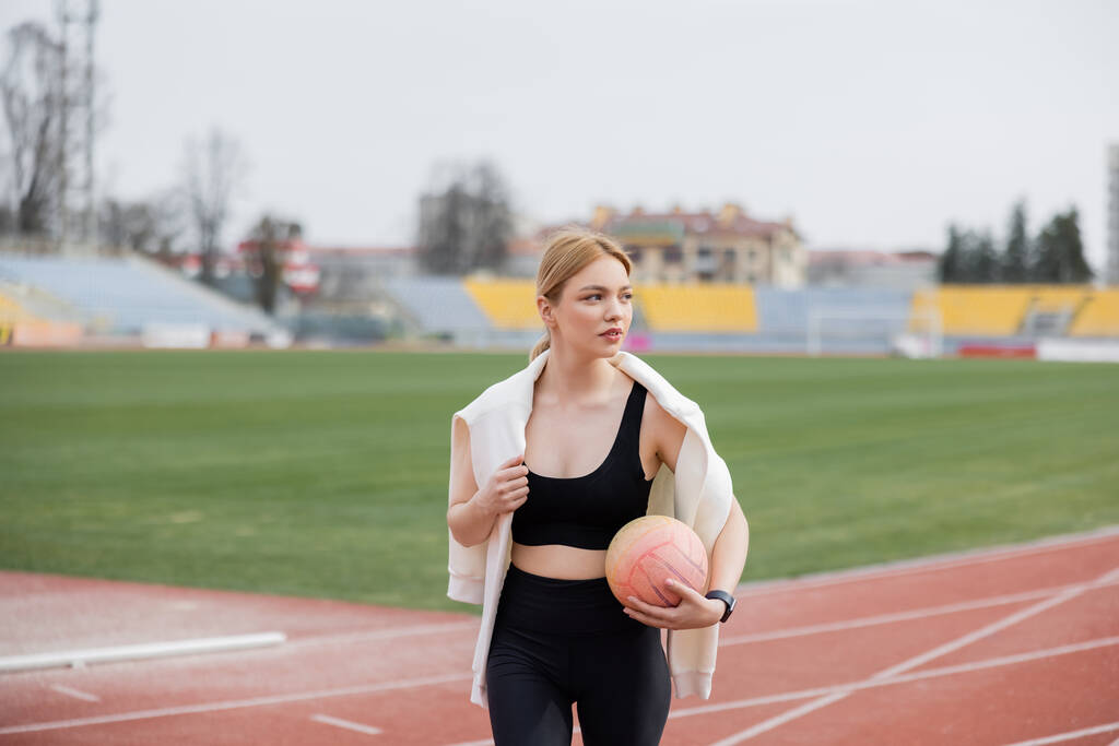 женщина в черной спортивной одежде стоит на стадионе с мячом и смотрит в сторону - Фото, изображение