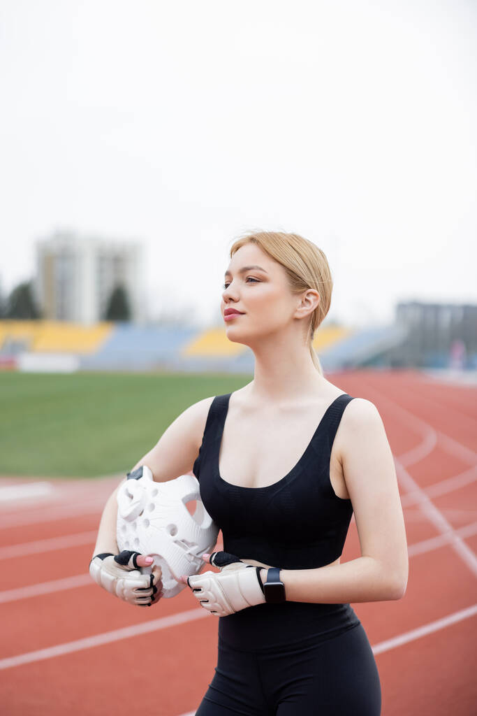 Αθλητική γυναίκα σε γάντια φυσικής κατάστασης κρατώντας αθλητικό κράνος και κοιτάζοντας μακριά σε εξωτερικούς χώρους - Φωτογραφία, εικόνα