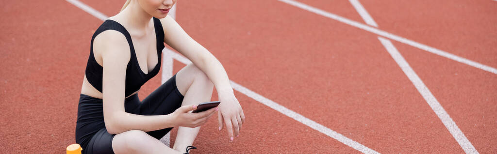 vista cortada de esportista sentado no campo atlético com telefone celular, banner - Foto, Imagem