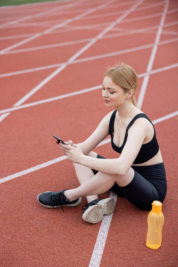 Sportliche Frau sitzt neben Sportflasche und benutzt Handy - Foto, Bild