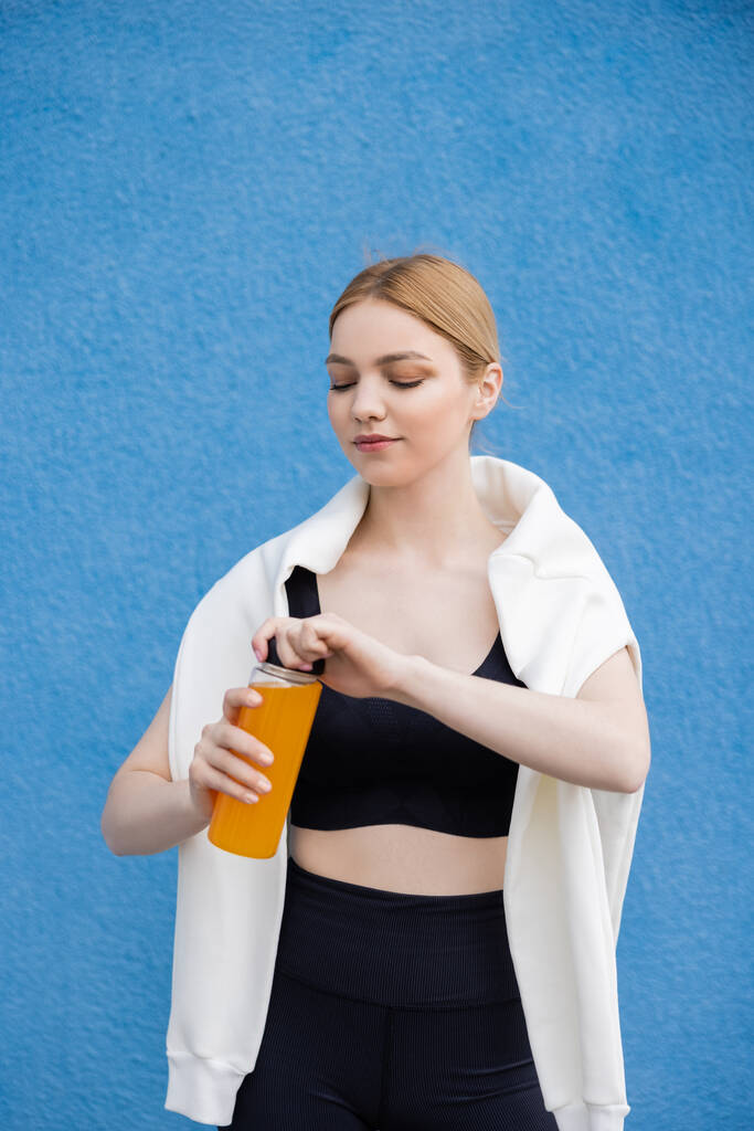 sportive woman in black sports bra opening bottle of fresh orange juice on blue textured background - Photo, Image