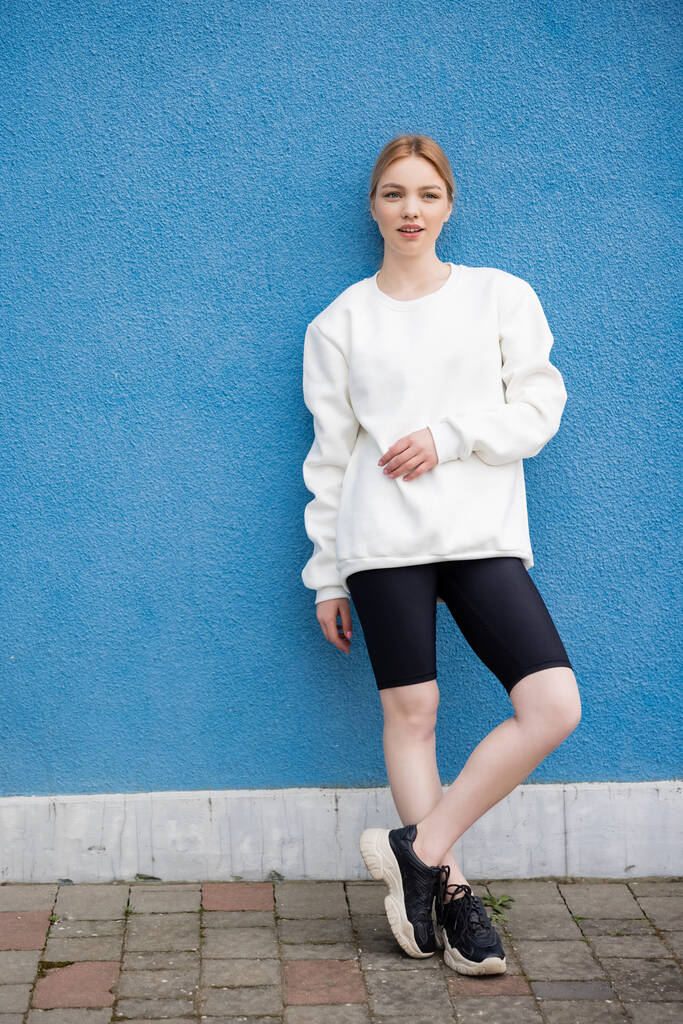 full length view of woman in white sweatshirt and black bike shorts near blue wall outdoors - Photo, Image