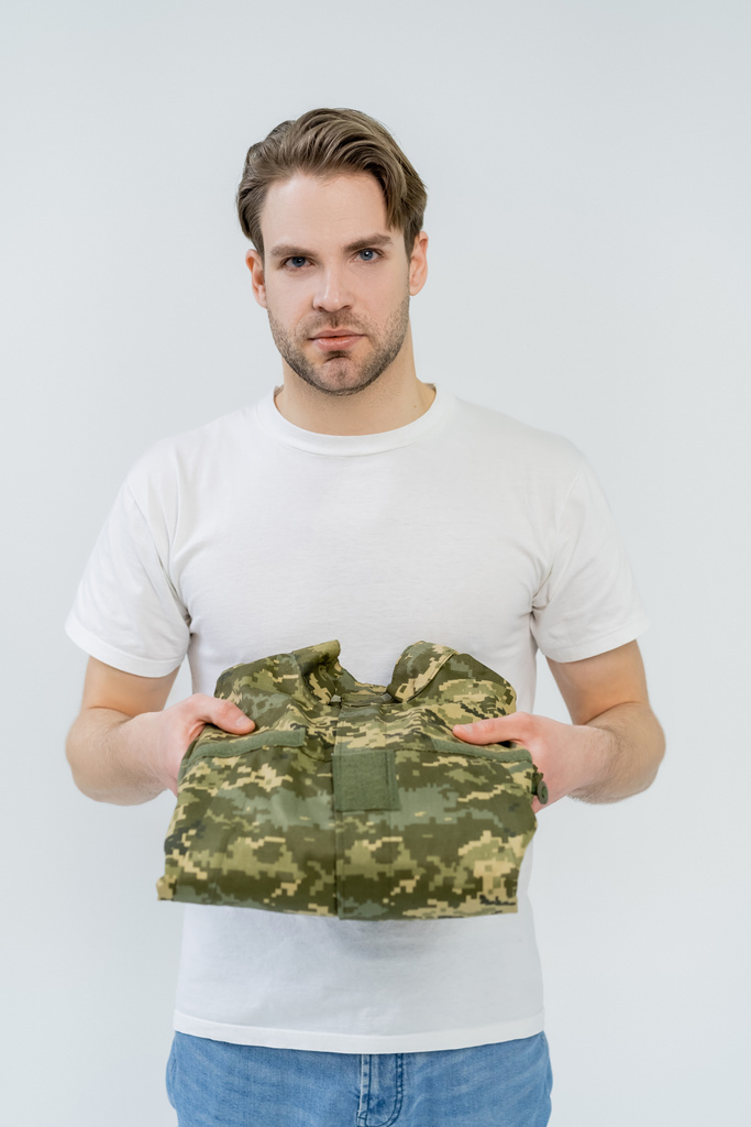 Young man holding military uniform and looking at camera isolated on white  - 写真・画像