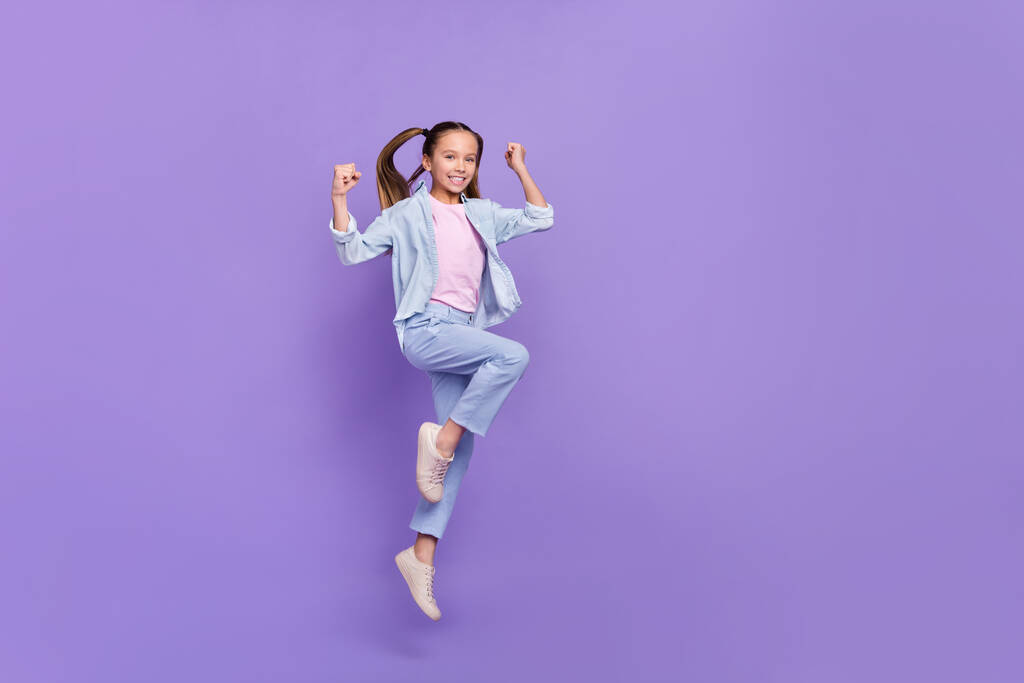 Kuva innoissaan melko pieni koulutyttö kulumista denim paita nousee nyrkit hyppäämällä suuri tyhjä tila eristetty violetti väri tausta - Valokuva, kuva