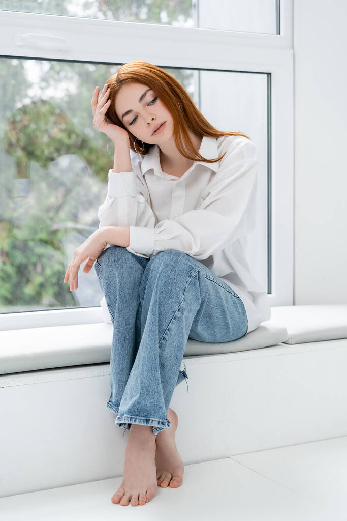 Barefoot vrouw in shirt en jeans zittend op vensterbank thuis - Foto, afbeelding