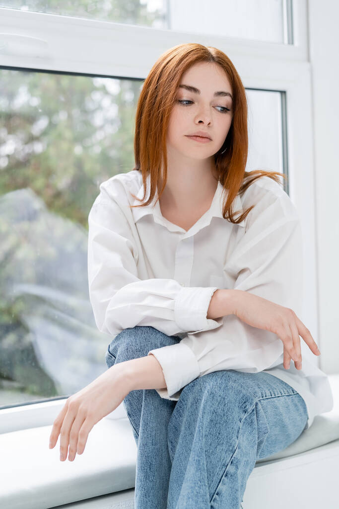 Järkyttynyt nainen paidassa istuu ikkunalaudalla kotona  - Valokuva, kuva
