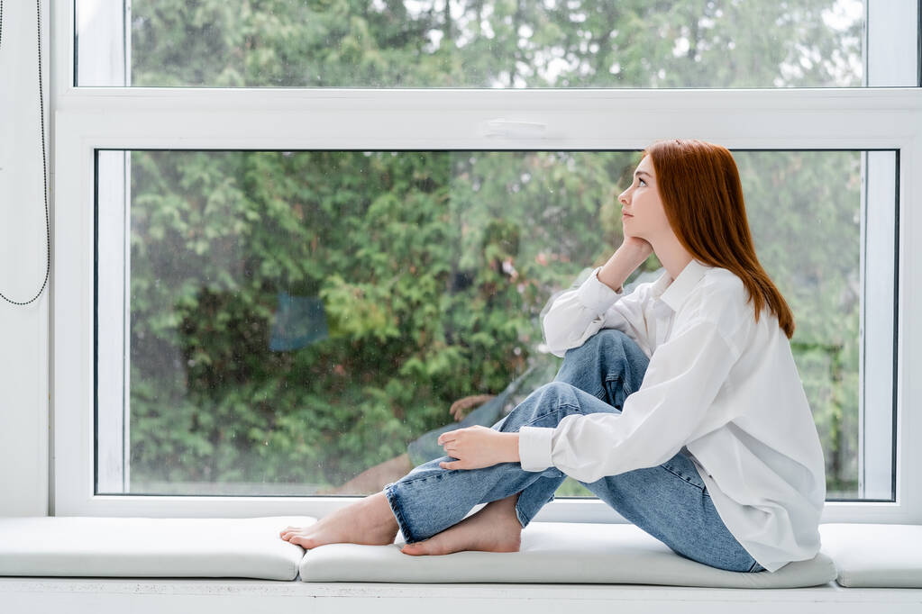 Widok z boku młoda ruda kobieta siedzi w pobliżu okna w domu  - Zdjęcie, obraz