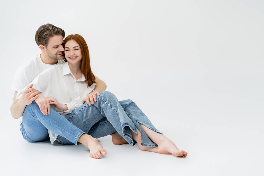 Smiling man in jeans kissing girlfriend in shirt on white background - Photo, Image