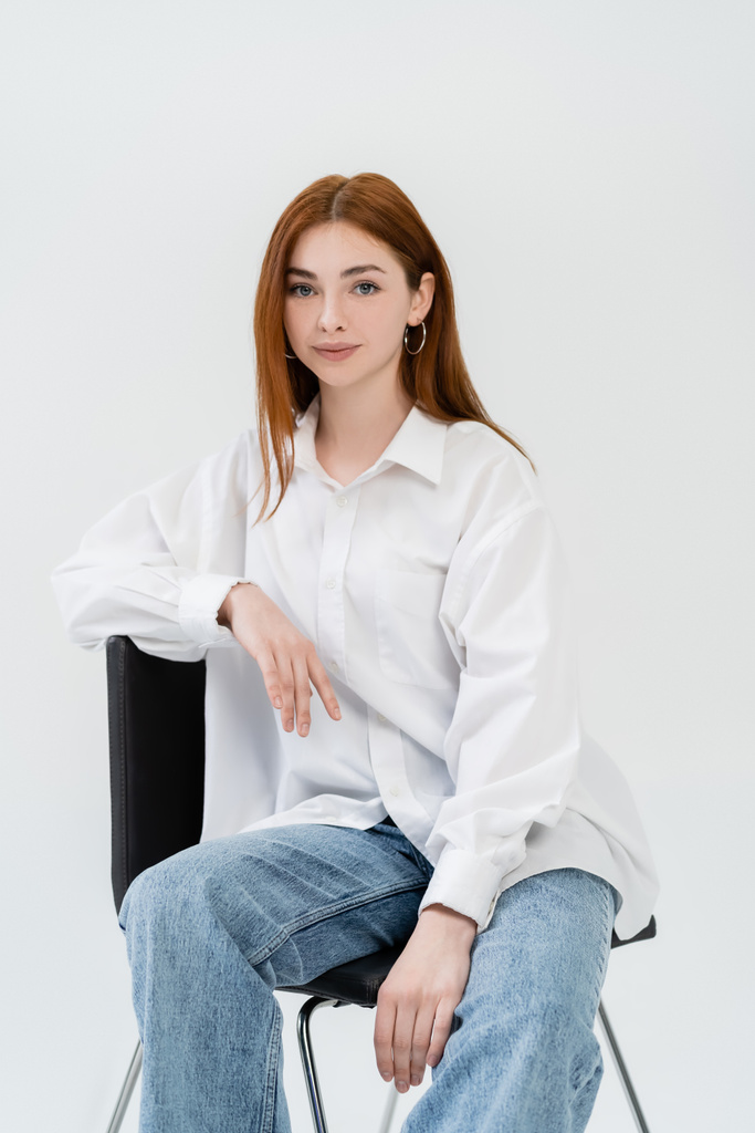 Redhead woman in shirt looking at camera while sitting on chair isolated on white  - 写真・画像