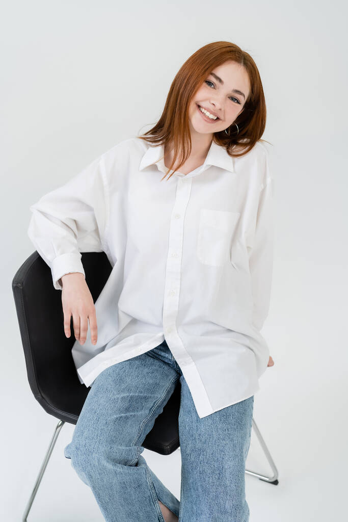 Iloinen nuori nainen paidassa katselee kameraa istuessaan tuolilla valkoisella taustalla - Valokuva, kuva