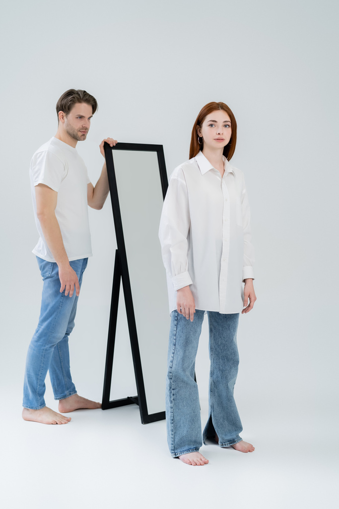 Pełna długość boso kobieta patrząc na aparat w pobliżu lustra i chłopaka na białym tle - Zdjęcie, obraz