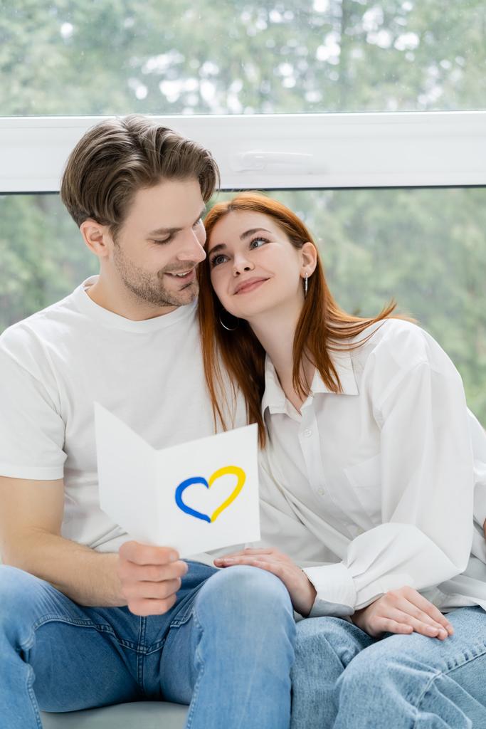 Positive man holding card with blue and yellow heart sign and looking at girlfriend at home - Photo, Image