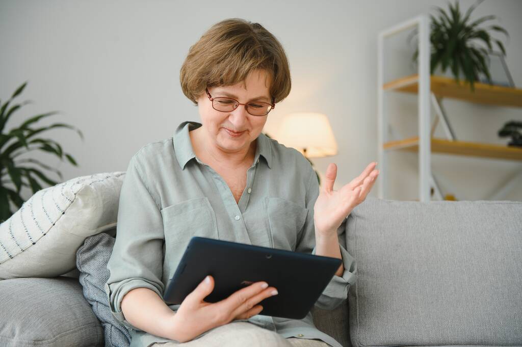 Seniorin auf dem Sofa in Wohnung oder Seniorenheim mit Tablet-Computer online. - Foto, Bild
