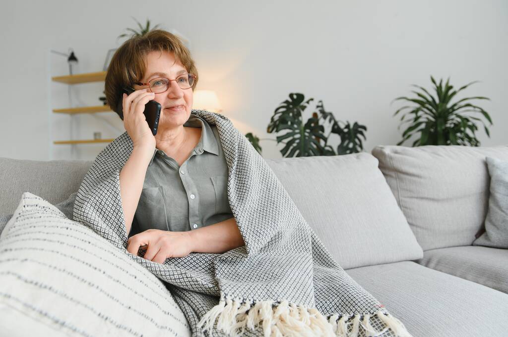 Красива старша жінка в повсякденному одязі, використовуючи свій смартфон, сидячи на дивані вдома
. - Фото, зображення