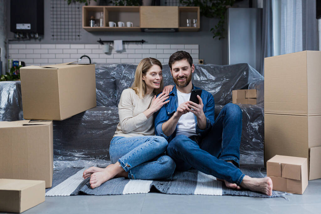 Ein Mann und eine Frau in einer neuen Wohnung, ein Ehepaar, das auf dem Boden sitzt, neben Pappkartons, das Telefon für Online-Einkäufe benutzt, Möbel auswählt - Foto, Bild