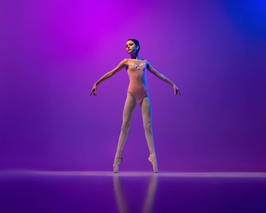 Studio shot van mooie school leeftijd meisje, ballet danser dansen geïsoleerd op paarse achtergrond in neon licht. Kunst, gratie, schoonheid, ballet school concept - Foto, afbeelding