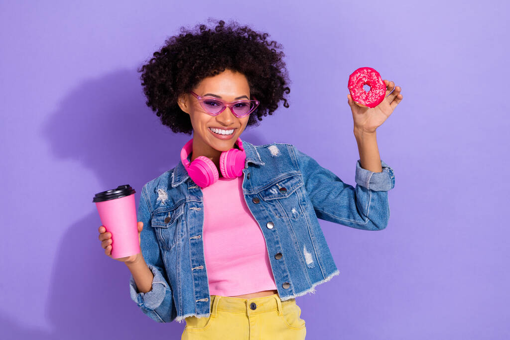 Фото мрійливої смішної молодої жінки в джинсовій куртці навушники, що п'ють чай, їдять печиво ізольований фіолетовий кольоровий фон
 - Фото, зображення