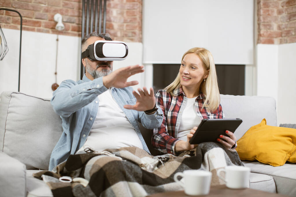 Hombre sentado en el sofá en los auriculares VR mientras que la mujer que usa la tableta - Foto, Imagen