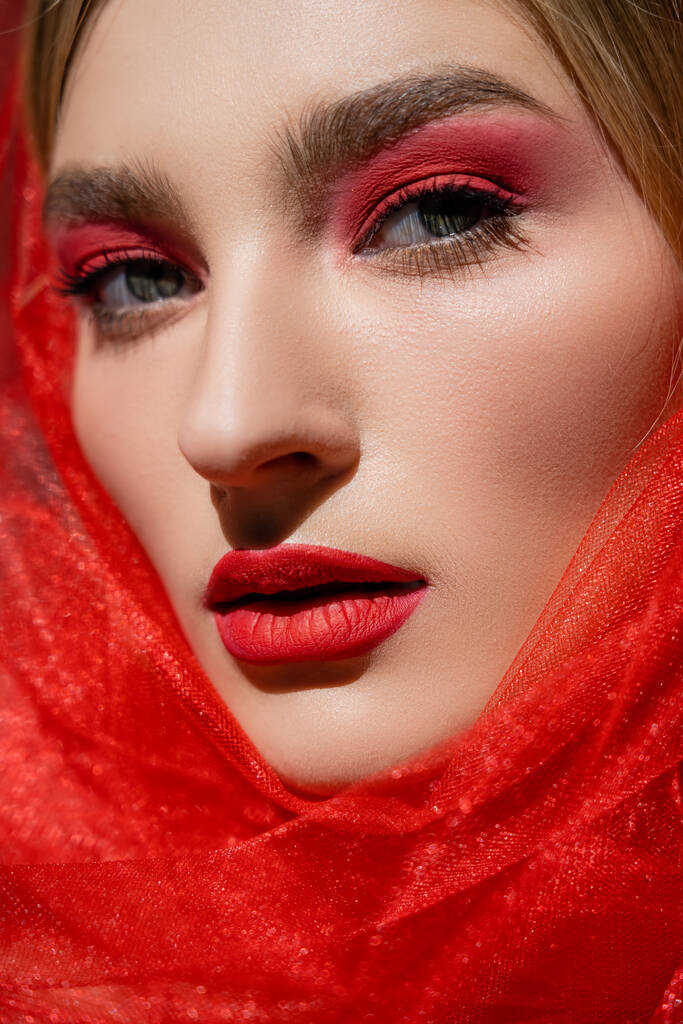 Close up view of young woman with red eye shadow and tulle cloth looking at camera  - Photo, Image