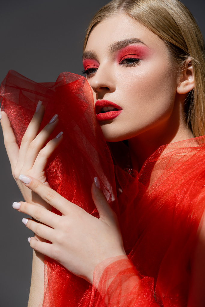 Όμορφη γυναίκα με μακιγιάζ κρατώντας κόκκινο τούλι ύφασμα απομονωμένο σε γκρι  - Φωτογραφία, εικόνα