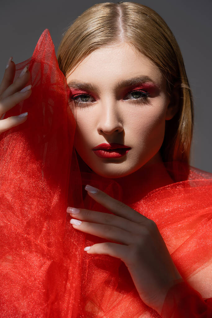 Portrait of young woman with red makeup holding tulle cloth and looking at camera isolated on grey  - Photo, Image