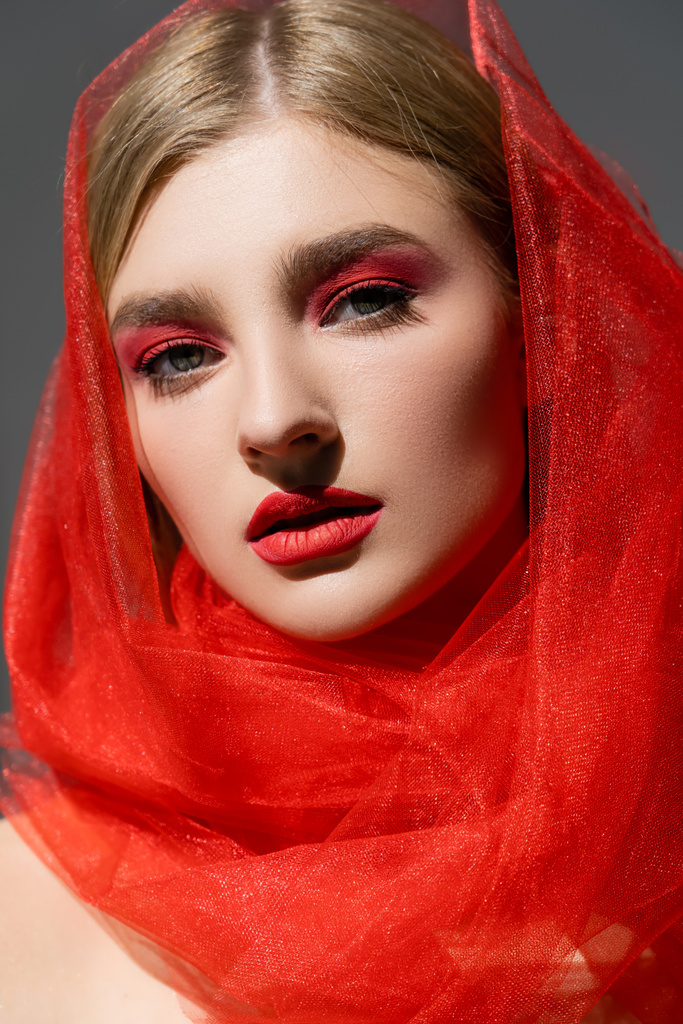 Retrato de modelo con rostro rojo y tela de tul mirando a la cámara aislada en gris  - Foto, Imagen