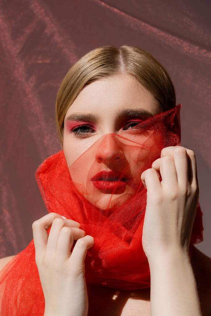 Fair haired model with red eye shadow touching tulle cloth on grey background  - 写真・画像