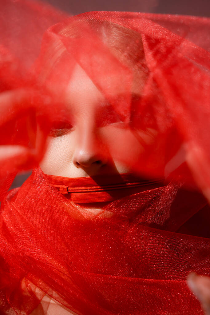 Junge Frau mit Reißverschluss am Mund und verschwommenem rotem Tuch auf grauem Hintergrund  - Foto, Bild