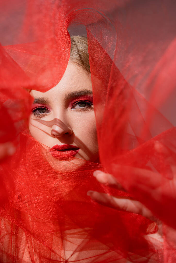 Молодая модель с красным макияжем, смотрящая на камеру возле размытой ткани на сером фоне  - Фото, изображение