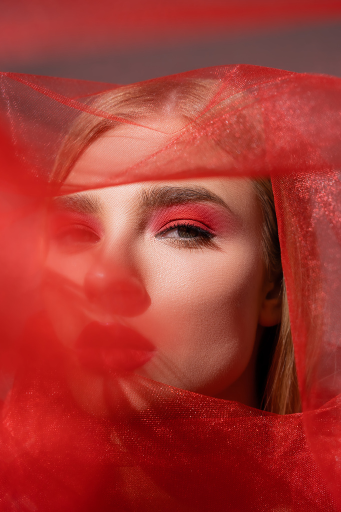 Μοντέλο με κόκκινη σκιά ματιών κοιτάζοντας κάμερα κοντά θολή πανί σε γκρι φόντο  - Φωτογραφία, εικόνα