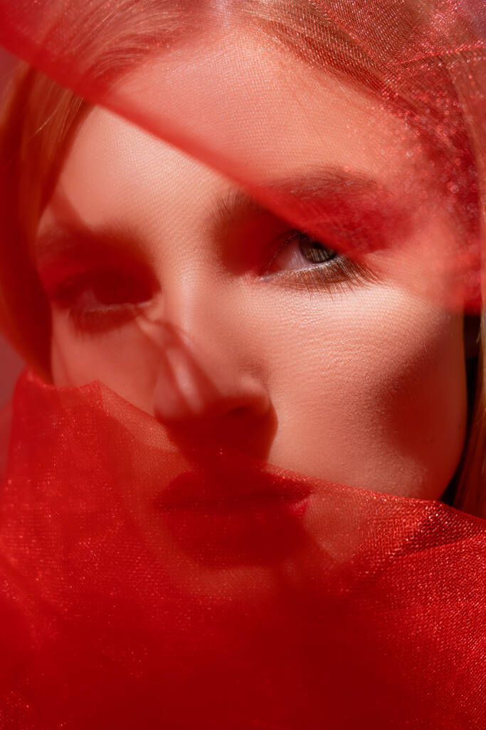 Close up view of young woman looking at camera near red blurred cloth  - Photo, Image
