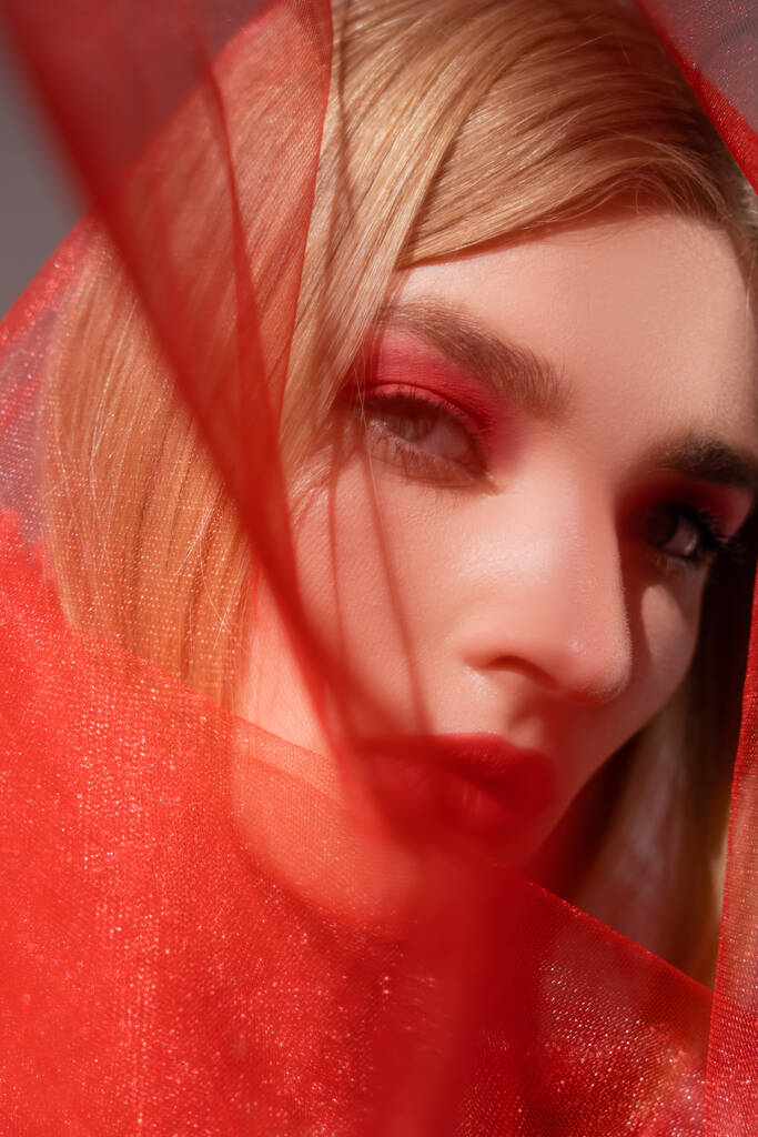 Θολή μοντέλο με κόκκινα χείλη κοιτάζοντας κάμερα κοντά σε ύφασμα σε γκρι φόντο  - Φωτογραφία, εικόνα
