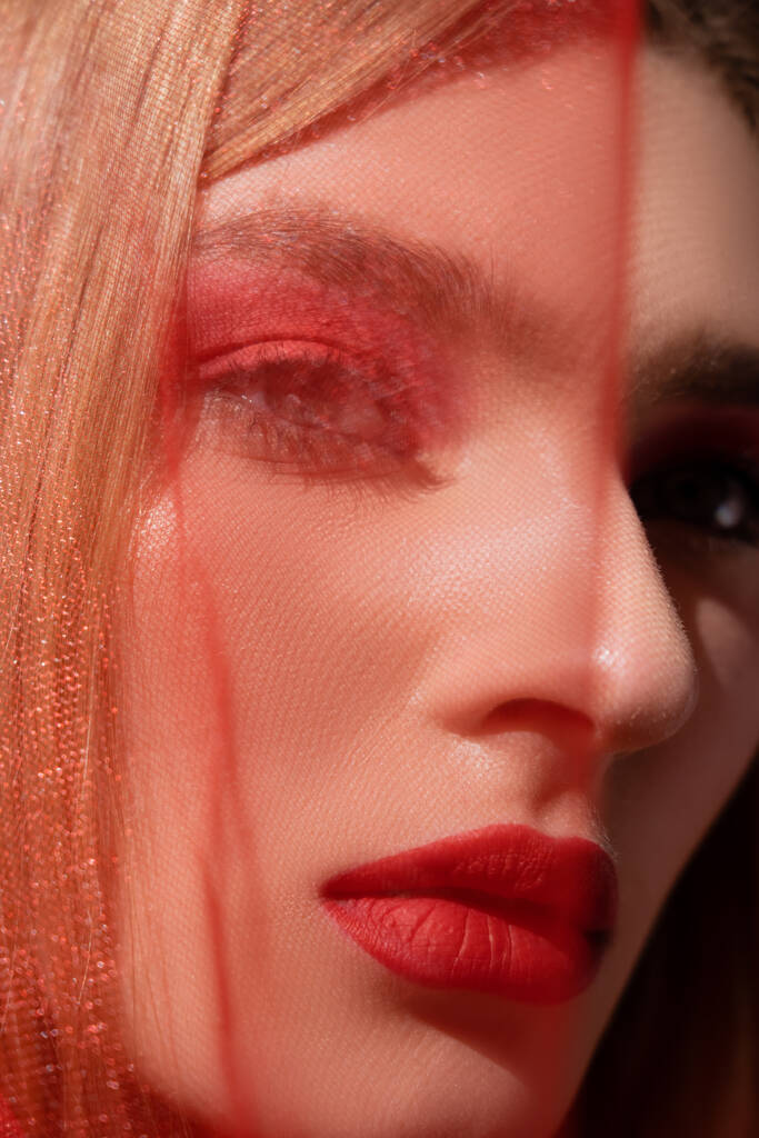 Close up widok kobiety z makijażem patrząc na aparat w pobliżu rozmytej czerwonej tkaniny izolowane na czarno  - Zdjęcie, obraz
