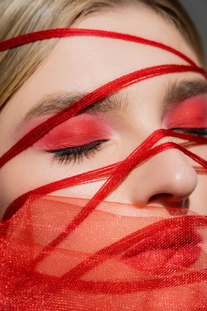Close up view of woman with red eye shadow, threads and fabric near face isolated on grey  - Photo, Image