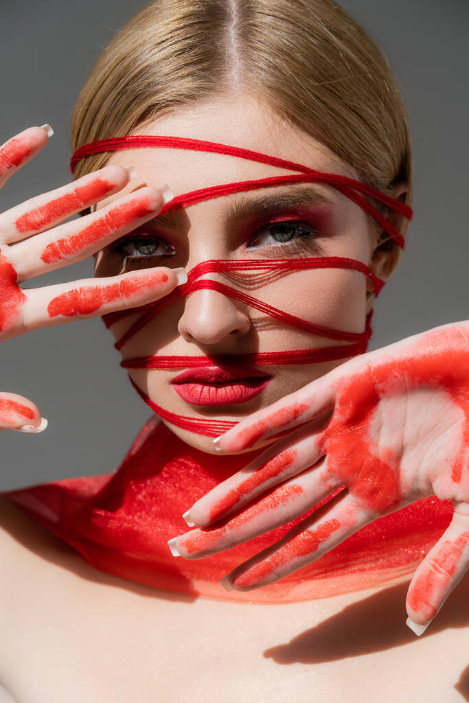 Jong model met rode draden op gezicht en verf op handen kijkend naar camera geïsoleerd op grijs  - Foto, afbeelding