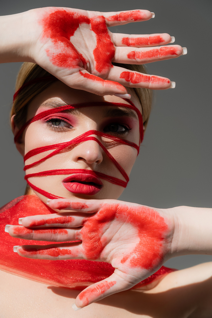 Junge Frau mit rotem Visage, Fäden und Farbe an den Händen posiert isoliert auf grau  - Foto, Bild