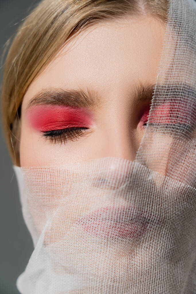 Close up view of woman with red eye shadow and medical bandage on face isolated on grey  - Фото, изображение