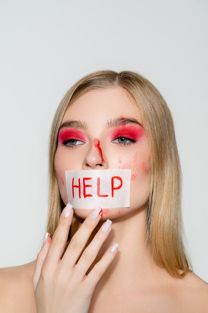 Mulher jovem com sangue no rosto e cartão com ajuda lettering em lábios isolados em cinza  - Foto, Imagem
