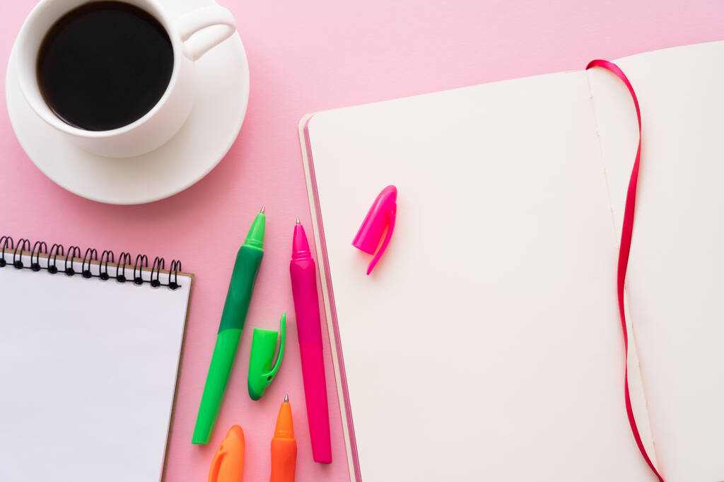 bovenaanzicht van kleurrijke pennen in de buurt van blanco notitieboekjes en kopje koffie op roze - Foto, afbeelding
