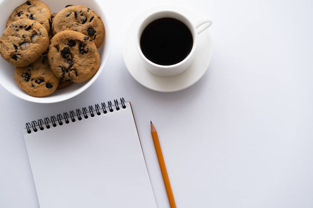 bovenaanzicht van kom met koekjes met chocolade chip in de buurt van kopje koffie en notebook op wit - Foto, afbeelding