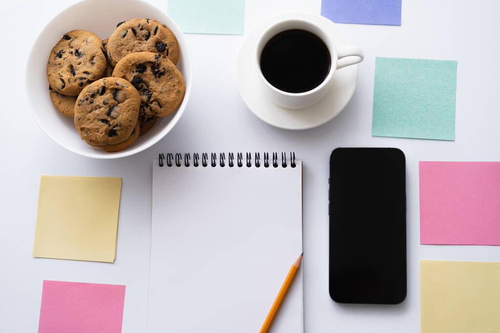top view of cup of coffee, cookies and stationery near smartphone with blank screen on white - Photo, image