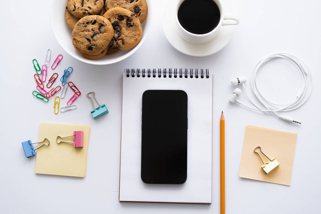 top view of cup of coffee, cookies and smartphone with blank screen near stationery on white - Фото, изображение