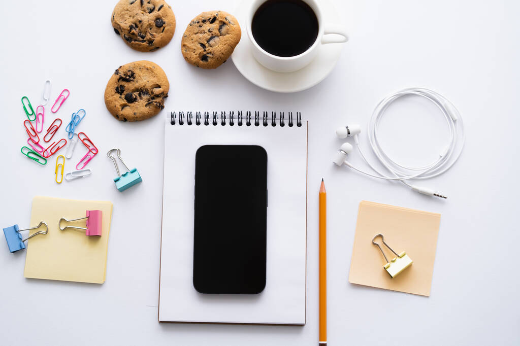 top view of cup of coffee, biscuits and smartphone with blank screen near stationery on white - Photo, Image