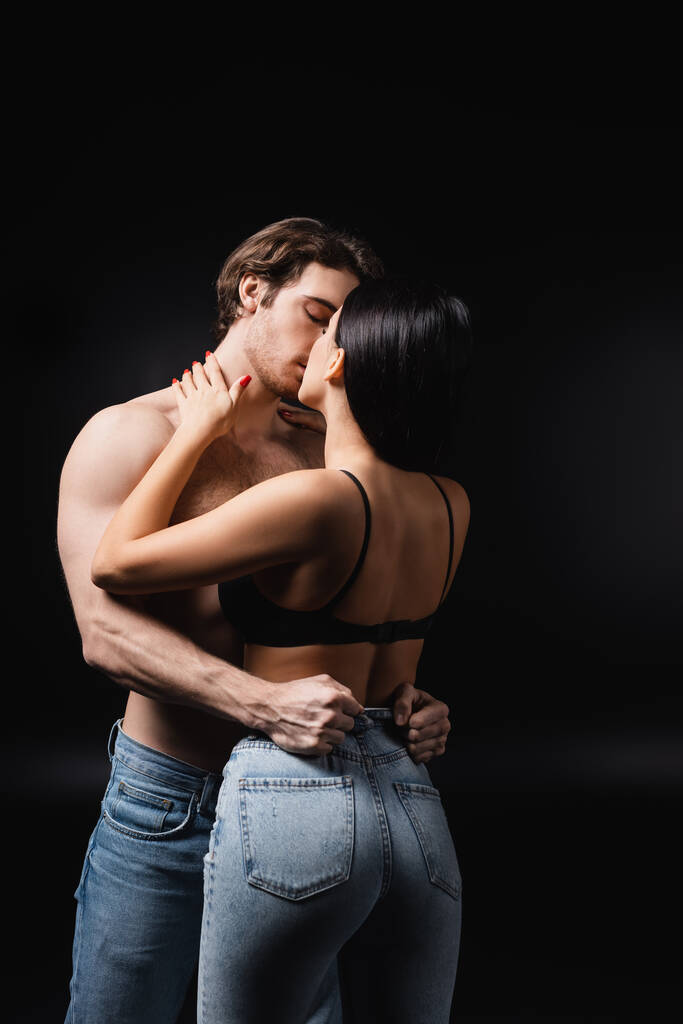 Woman in jeans and bra kissing sexy boyfriend isolated on black  - Foto, Imagem