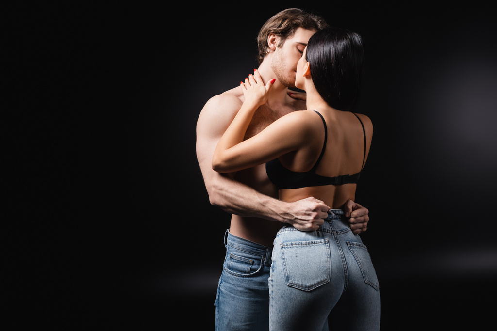 Страстная брюнетка целует мужчину без рубашки на черном фоне - Фото, изображение
